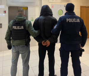 Na korytarzy tyłem stoi trzech mężczyzn. Umundurowany policjant trzyma pod rękę mężczyznę, który ma założone kajdanki na ręce trzymane z tyłu. Z drugiej strony zatrzymanego mężczyzny stoi policjant, który na sobie ma kamizelkę kuloodporna z napisem POLICJA i tak samo trzyma pod pachę zatrzymanego.