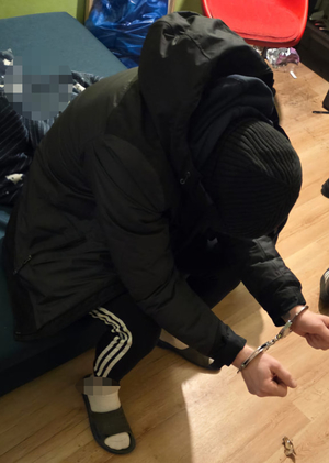 W pokoju na łóżku siedzi pochylony mężczyzna. Ubrany jest w czarną bluzę z kapturem i czarną kurtkę z kapturem. Na głowie ma założoną czarną czapkę. Ma tez czarne dresowe spodnie z trzema białymi paskami. Mężczyzna ręce ma wyciągnięte do przodu, na których ma założone kajdanki. W pokoju jest bałagan.