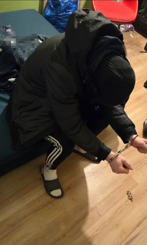 W pokoju na łóżku siedzi pochylony mężczyzna. Ubrany jest w czarną bluzę z kapturem i czarną kurtkę z kapturem. Na głowie ma założoną czarną czapkę. Ma tez czarne dresowe spodnie z trzema białymi paskami. Mężczyzna ręce ma wyciągnięte do przodu, na których ma założone kajdanki. W pokoju jest bałagan.