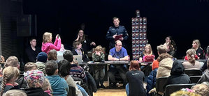 Na sali na krzesełkach siedzi duża grupa osób w większości to młodzież. Patrzą na osoby, które siedzą na przeciwko nich przy podłużnym stoliku. Za nimi stoi piramida z puszek WOŚP, obok której stoi z mikrofonem umundurowany policjant.