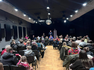 Na sali na krzesełkach siedzi duża grupa osób w większości to młodzież. Patrzą na osoby, które siedzą na przeciwko nich przy podłużnym stoliku. Za nimi stoi piramida z puszek WOŚP, obok której stoi z mikrofonem umundurowany policjant.