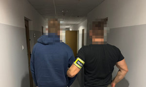 zatrzymany mężczyzna w asyście nieumundurowanego policjanta, obywaj stoją tyłem. Policjant  ma na ręku odblaskową opaskę z napisem Policja, zdjęcie wykonane wewnątrz budynku, na korytarzu