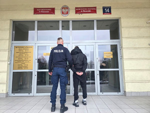 zatrzymany mężczyzna w asyście nieumundurowanego policjantka, w tle wejście do budynku Prokuratury Rejonowej w Piasecznie
