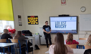 policjantka stojąca przy ekranie multimedialnym, na którym widnieje napis "Lód jest kruchy". Dzieci siedzą w ławkach szkolnych