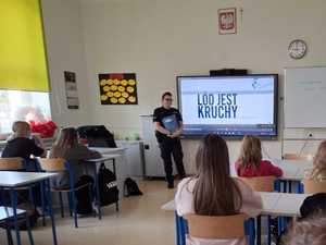 policjantka stojąca przy ekranie multimedialnym, na którym widnieje napis "Lód jest kruchy". Dzieci siedzą w ławkach szkolnych