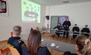 zdjęcie wykonane w trakcie debaty społecznej, widać na nim siedzących przy stole mieszkańców, w tle policjantów, a na wyświetlaczu plakat promujący plebiscyt #SuperDzielnicowy