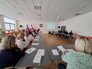 zdjęcie wykonane w trakcie debaty społecznej, widać na nim siedzących przy stole mieszkańców, w tle policjantów, a na wyświetlaczu slajd z przygotowanej prezentacji