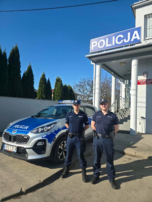 umundurowani policjanci stojący przy radiowozie, w tle budynek Komisariatu Policji w Górze Kalwarii