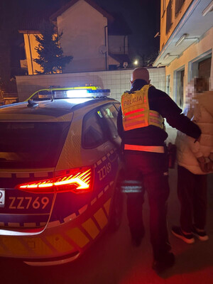 umundurowany policjantów wraz z zatrzymaną kobietą, która ma założone kajdanki na ręce trzymane z tyłu. W tle policyjny oznakowany radiowóz