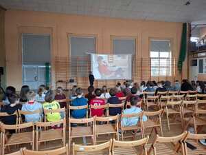 policjantka stojąca obok projektora, młodzież siedząca na krzesłach. Zajęcia odbywały się na hali sportowej