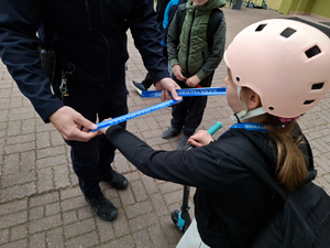 Umundurowany policjant zakłada na rękę dziewczynki pasek odblaskowy. Dziewczynka ma na głowie kask do jazdy na hulajnodze.