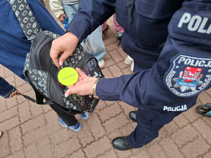 Umundurowana policjantka przyczepia plecaka szkolnego element odblaskowy. Plecak ma na sobie uczeń.