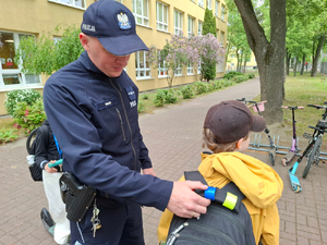 Umundurowany policjant przyczepia do plecaka szkolnego element odblaskowy.