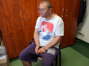 Na krzesełku siedzi mężczyzna ubrany w bluzkę z krótkim rękawem i w krótkich spodenkach. Na rękach trzymanych z przodu ma założone kajdanki.