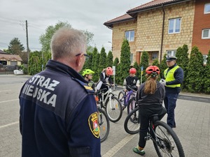 policjant wydziału ruchu drogowego rozmawiający z dziećmi, w tle strażnik miejski