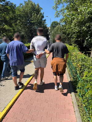 zatrzymany mężczyzna w asyście nieumundurowanych policjantów, zatrzymany ma założone kajdanki na ręce trzymane z tyłu