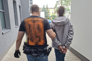 Policjant ubrany po cywilnemu prowadzi zatrzymanego mężczyznę, który ma założone kajdanki na ręce trzymane z tyłu.