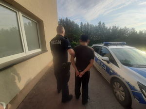 Umundurowany policjant prowadzi zatrzymanego mężczyznę , który ma założone kajdanki na ręce trzymane z tyłu.