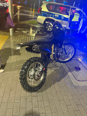 Na chodniku stoi czarny motocykl typu cross, a obok niego oznakowany radiowóz policyjny i umundurowany policjant.