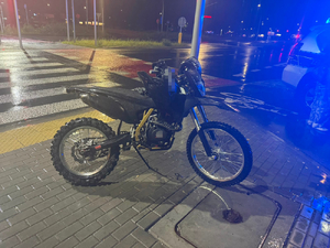 Na chodniku stoi czarny motocykl typu cross, a obok niego oznakowany radiowóz policyjny.