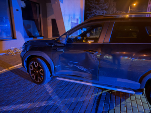 Na chodniku stoi ciemne auto, które ma wgnieciony bok od kierowcy.