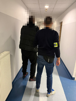 zatrzymany mężczyzna w asyście nieumundurowanego policjanta, który ma opaskę odblaskową z napisem "POLICJA" założoną na rękę