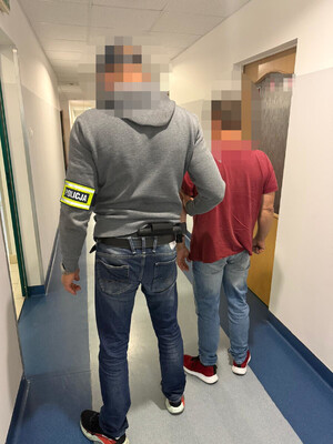 zatrzymany mężczyzna w asyście nieumundurowanego policjanta, który ma opaskę odblaskową z napisem "POLICJA" założoną na rękę