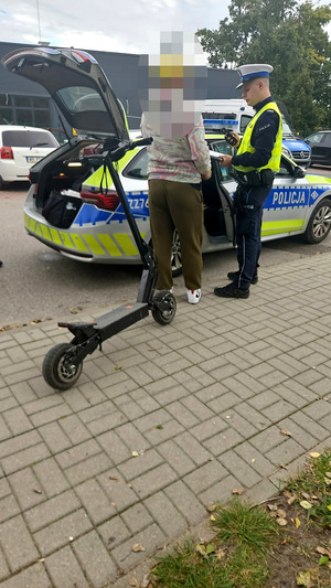 hulajnoga elektryczna, a w tle policyjny radiowóz obok którego stoi umundurowany policjant wydziału ruchu drogowego, jak również opiekun prawny, który zgłosił się po 12-latka zatrzymanego do kontroli drogowej