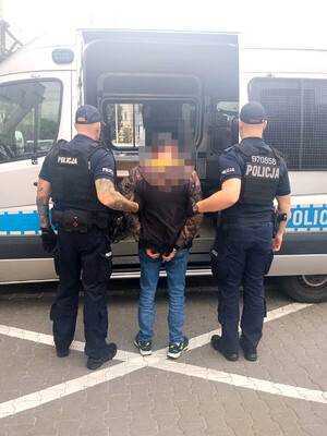 umundurowani policjanci razem z zatrzymanym mężczyzną, który ma założone kajdanki na ręce trzymane z tyłu. W tle policyjny oznakowany radiowóz