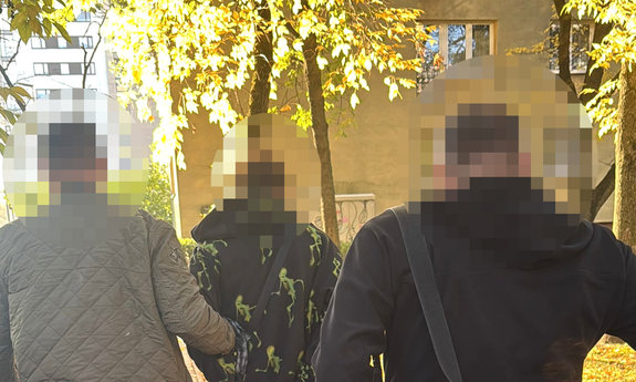 zatrzymany mężczyzna z kajdankami na rękach, w asyście nieumundurowanych policjantów. W tle blok mieszkalny i jesienne drzewa