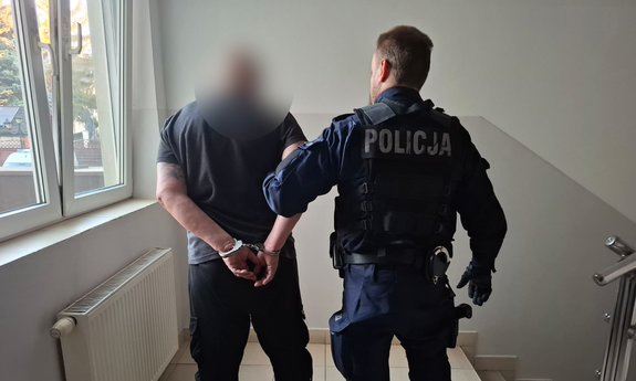 zatrzymany mężczyzna z kajdankami założonymi na ręce trzymane z tyłu, obok umundurowany policjant, w tle korytarz