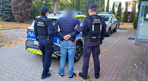 zatrzymany mężczyzna w asyście umundurowanych policjantów, w tle policyjny radiowóz