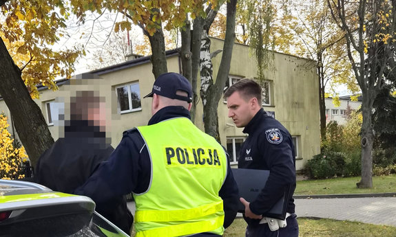 zatrzymany mężczyzna w asyście umundurowanych policjantów, w tle Komisariat Policji w Lesznowoli